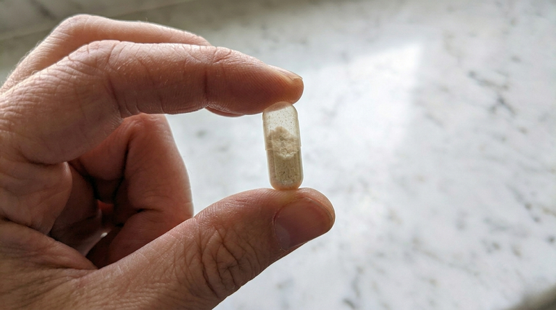 Close-up of fingers holding a single clear capsule filled with white calcium pyruvate powder against a white marble background
